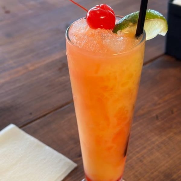 Tall glass of orange and red drink with crushed ice, a cherry, lime wedge, and black straw.