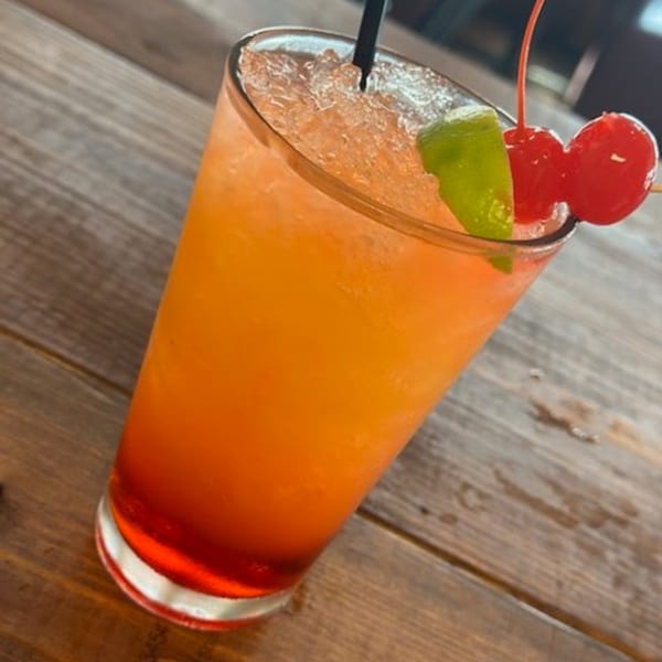 A vibrant orange and red cocktail, garnished with a lime wedge and two cherries, sits on a wooden table.