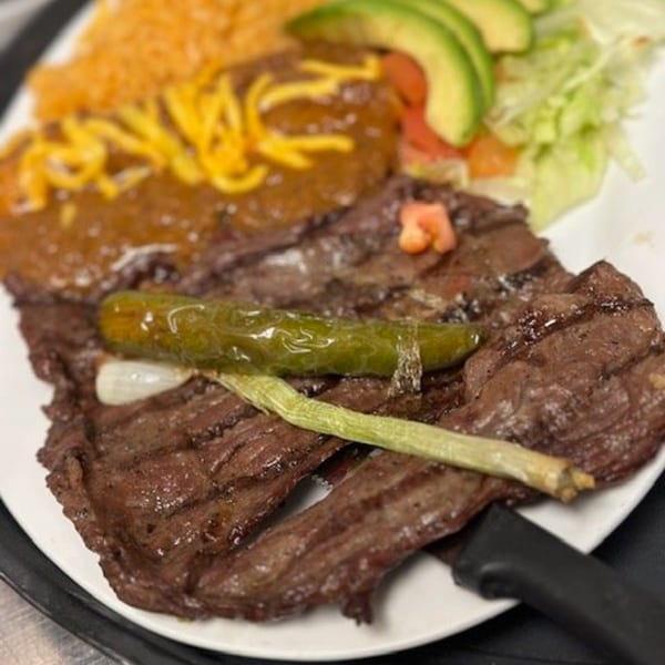 Grilled steak with a green chili, rice, beans, avocado, and salad on a white plate.