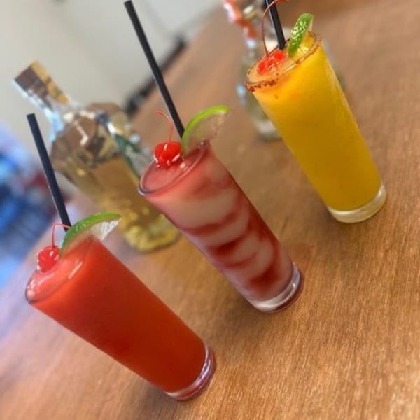 Three colorful frozen drinks, garnished with cherries and lime, on a wooden table at Los Charros.