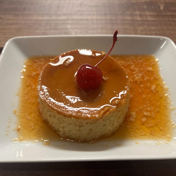 A round flan dessert with caramel sauce and a single red cherry on top, served on a white plate.