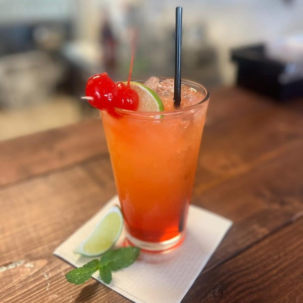 A vibrant orange and red cocktail, garnished with cherries, lime, and a mint sprig on a wooden table.