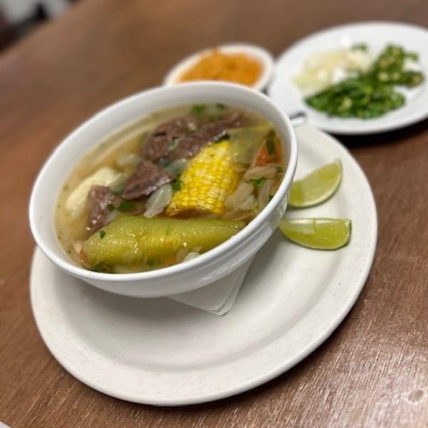 Hearty Mexican beef soup with corn, zucchini, and lime wedges, served with sides of chopped onions and jalapeños.