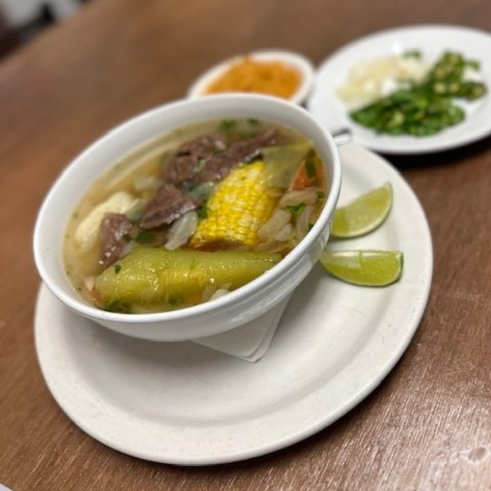 Hearty Mexican beef soup with corn, zucchini, and lime wedges, served with sides of chopped onions and jalapeños.