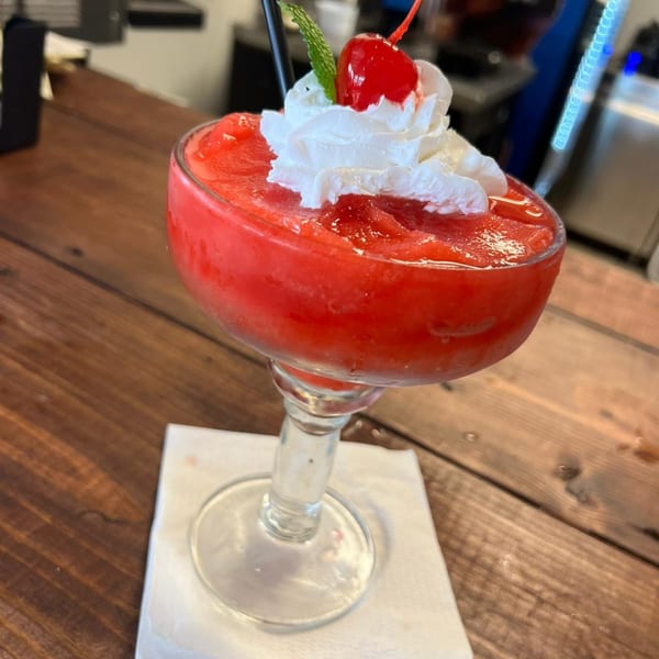A vibrant red frozen margarita, topped with whipped cream, a cherry, and mint, on a wooden table.
