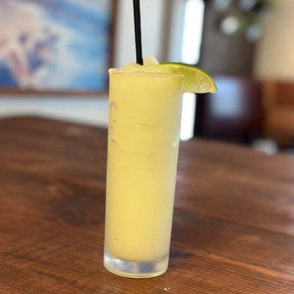 A frosty margarita with a salted rim, lime wedge, and black straw on a wooden table.