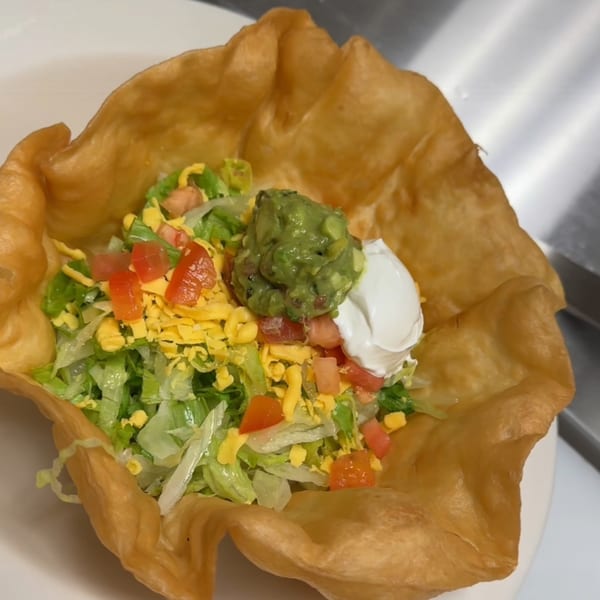 A taco salad in a crispy fried tortilla bowl, topped with lettuce, cheese, tomatoes, guacamole, and sour cream.